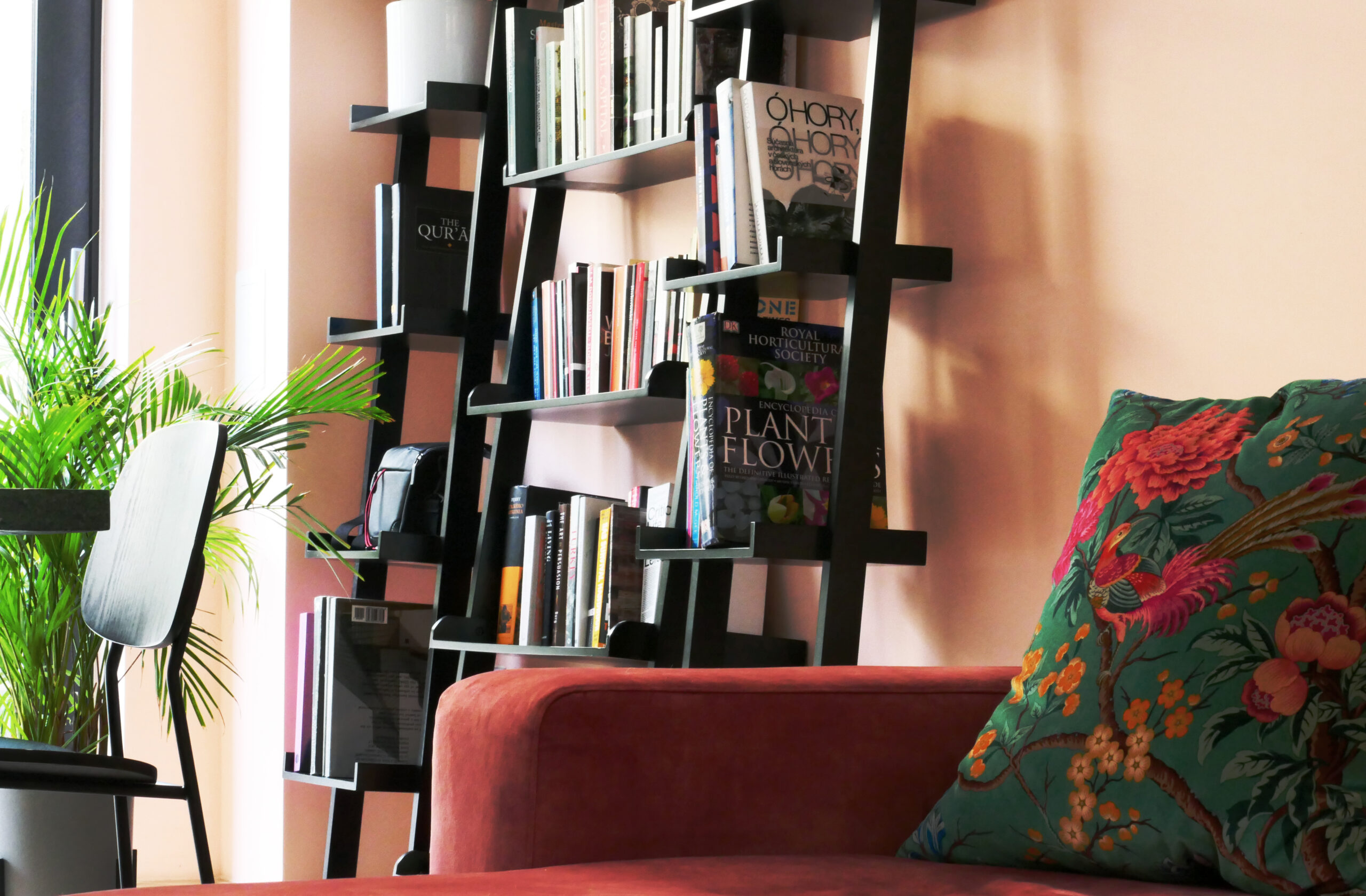 Cozy interior showing a sofa and a pillow with floral pattern. There is a plant and a bookshelf behind.